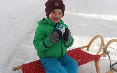 Lachendes Kind sitzt auf Schlitten im Schnee, Winterspaß beim Familienurlaub am Kinderbauernhof